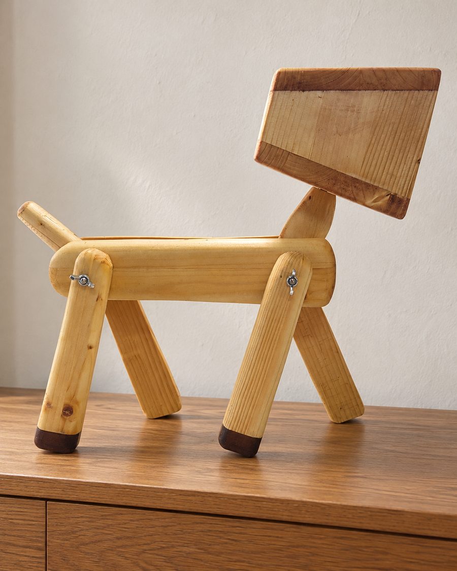 Un cane in legno chiaro con le zampe snodate e la testa a forma di scatola, su una credenza di rovere davanti a un muro chiaro