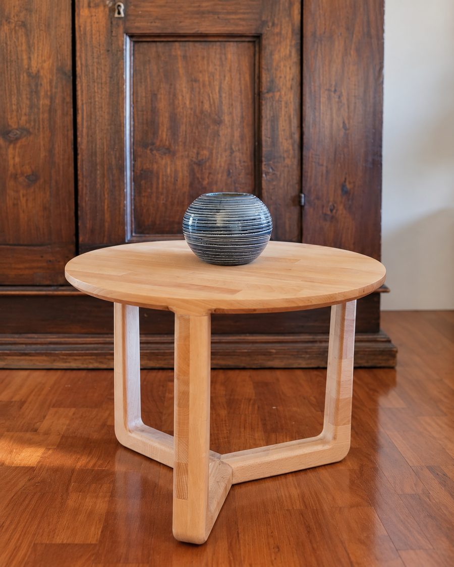 Un tavolino rotondo in legno chiaro con le gambe incrociate, davanti a un vecchio armadio di legno scuro